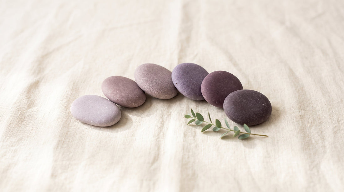 Six smooth stones in muted lavender and plum tones arranged in an arc on cream linen